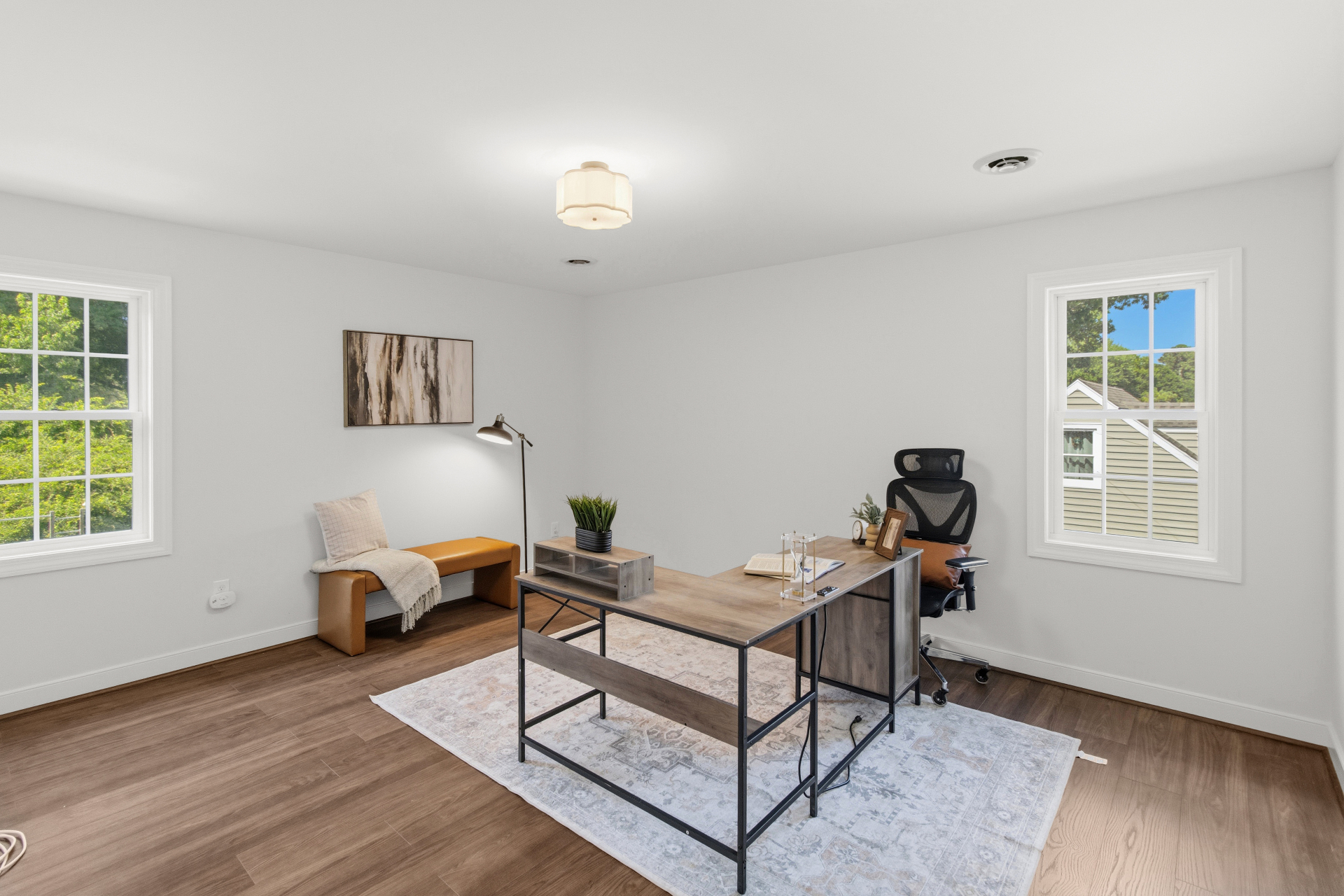 Staged home office with hardwood floors, neutral rug, leather bench, and L-shaped desk