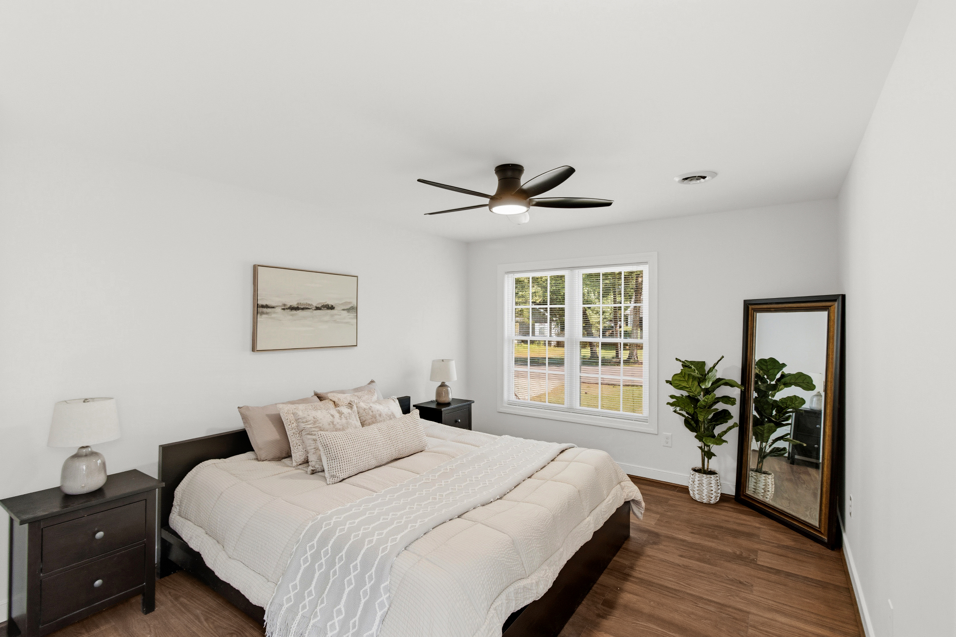 Staged bedroom with layered neutral bedding, dark nightstands, and wide front-facing windows